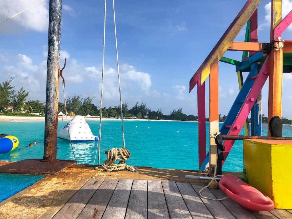 A dock overlooking Carlisle Bay in Bridgetown Barbados. In the foreground there is a platform painted in rainbow colors and in the background there are inflatable water toys