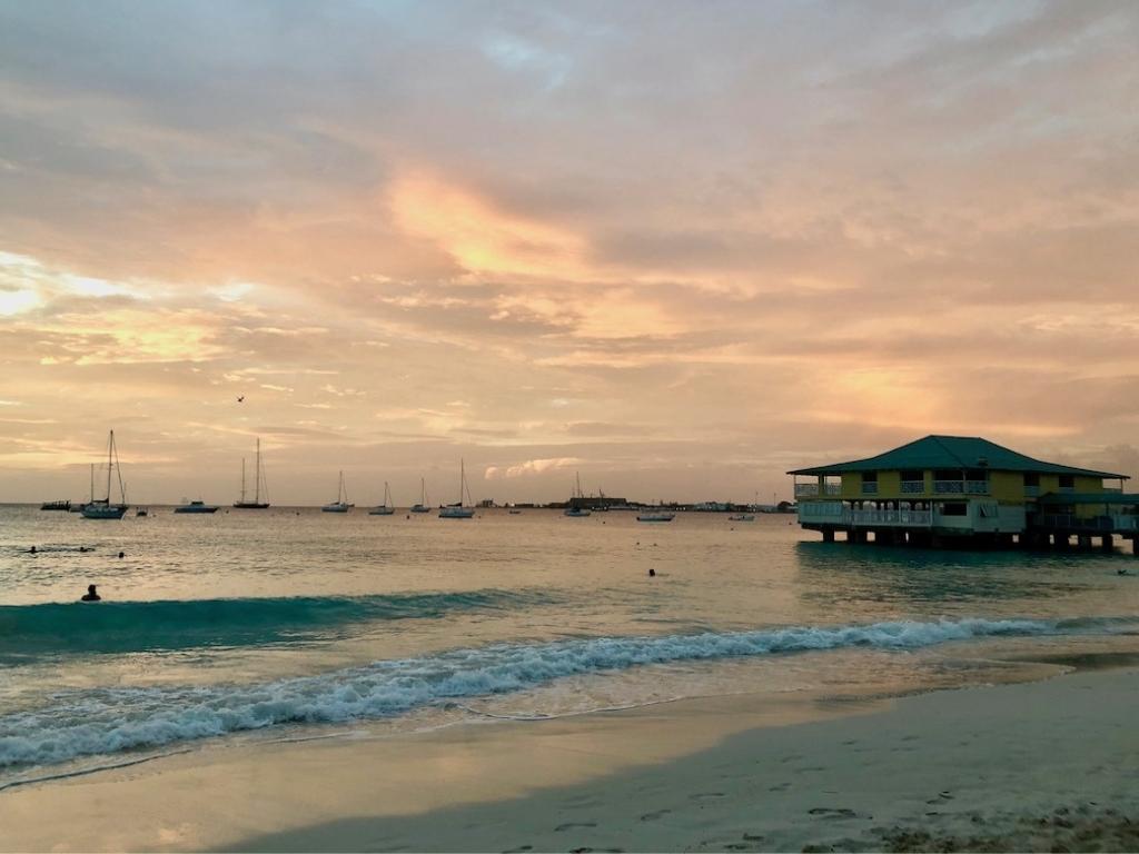 Pink and orange sunset at Pebbles Beach Barbados. There are sailboats in the background and a few people swimming in the foreground