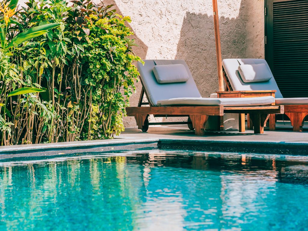 A blue pool lined in green plants with two lounge chairs under an umbrella