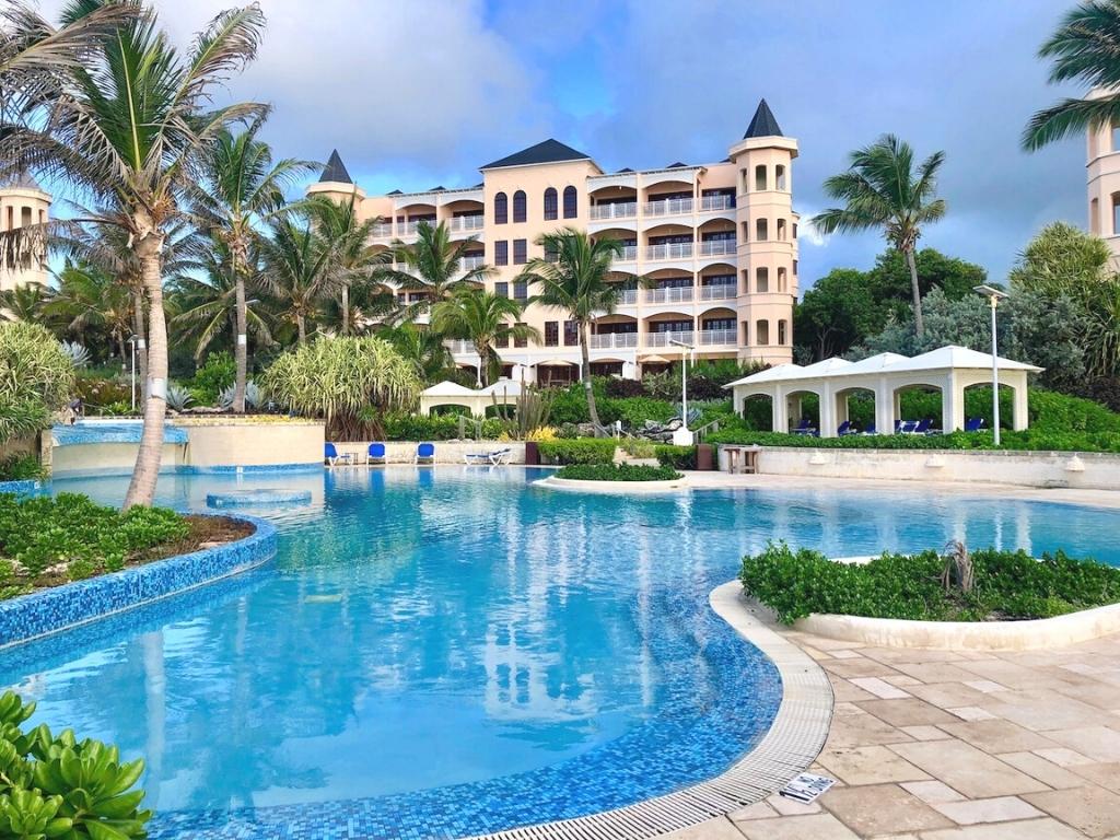 The pool area at the Crane Resort in Barbados. In the foreground you see a rounded pool surrounded by palm trees and in the background you can see a pink hotel with balconies and towers